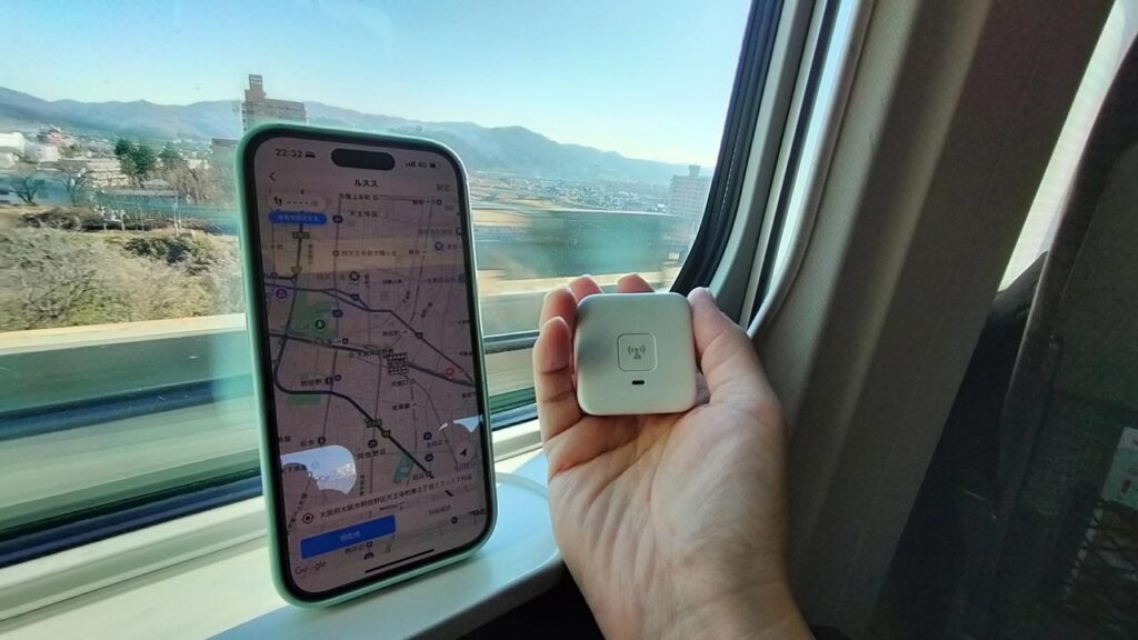 新幹線の車窓とスマホを一緒に写し、複数のこども用GPSアプリのログを見比べながら精度を検証している様子。移動中の位置ズレを確認しているイメージ写真。